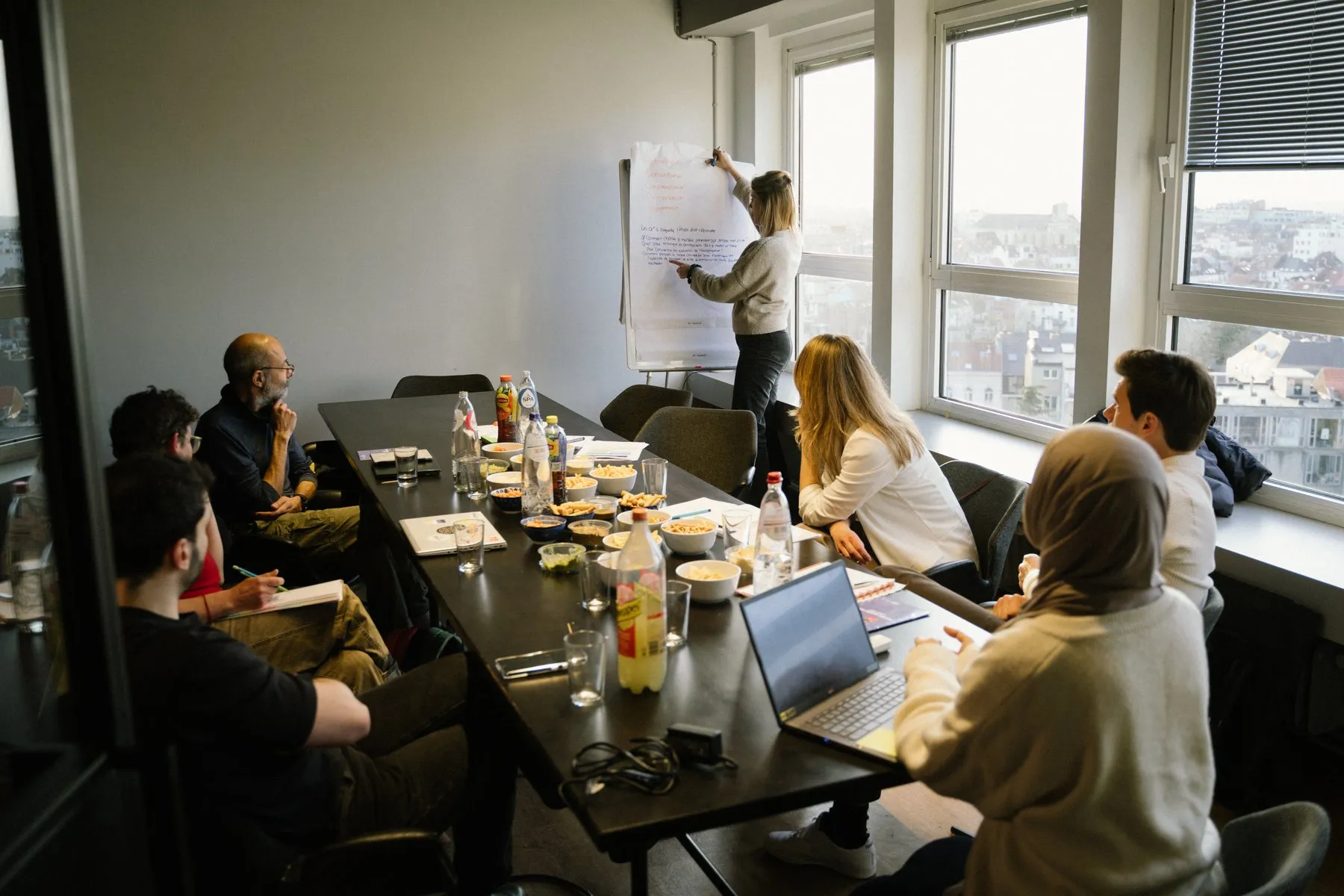 A working session around one long table in Brussels, notes on a flipchart and the city through the window.