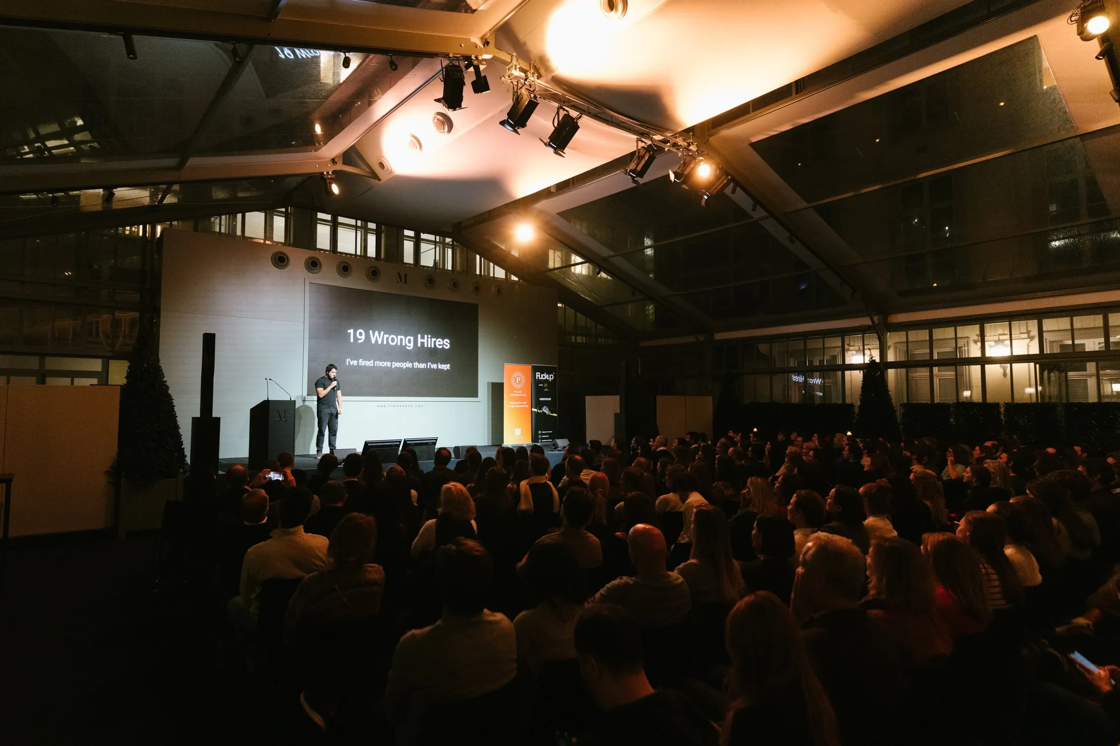 A dark hall under a glass roof, a packed audience, and a founder on stage beside a slide that reads nineteen wrong hires.