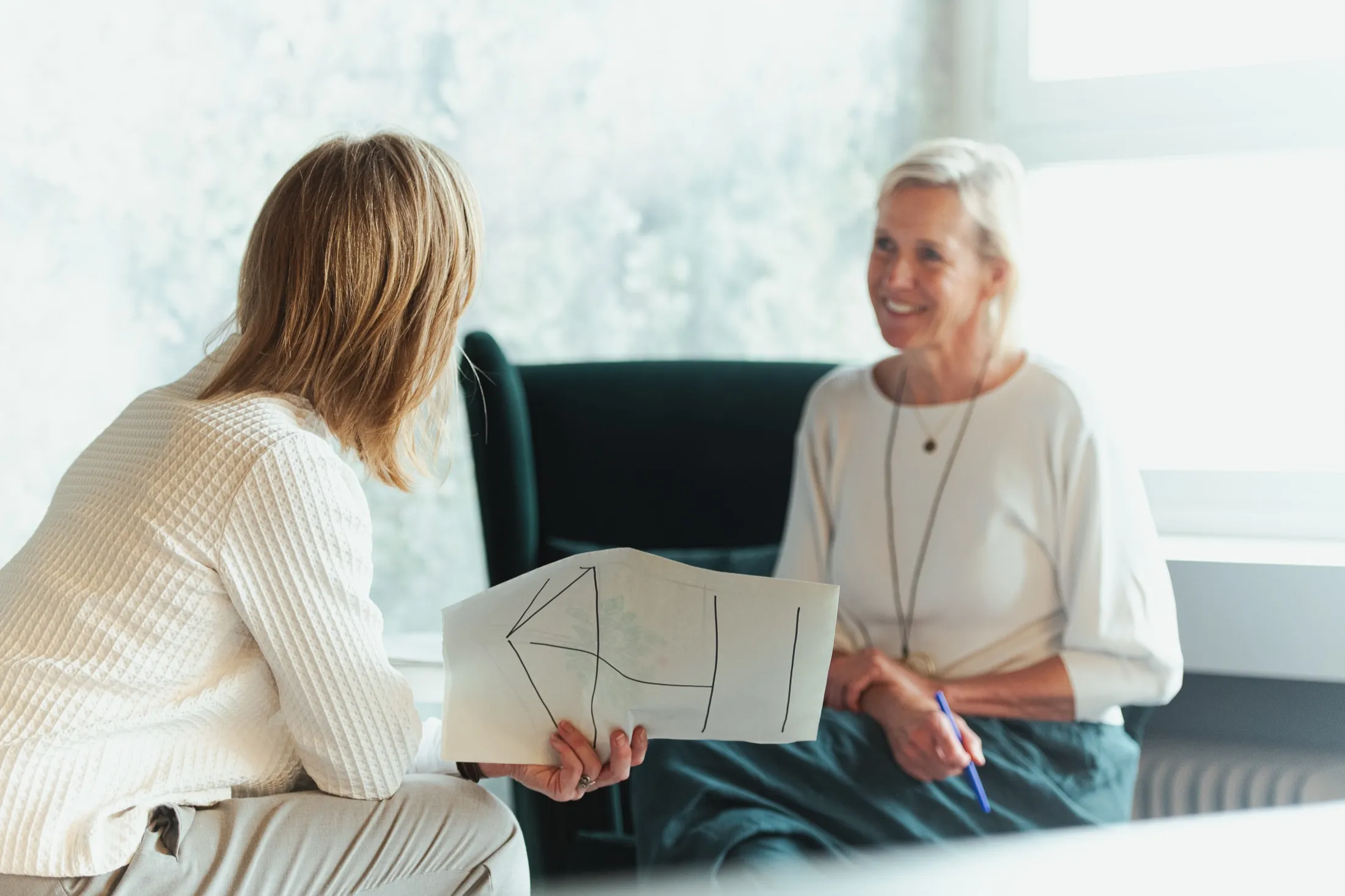 Two women in conversation at a Revival session, one holding a hand-drawn plan, in bright daylight.